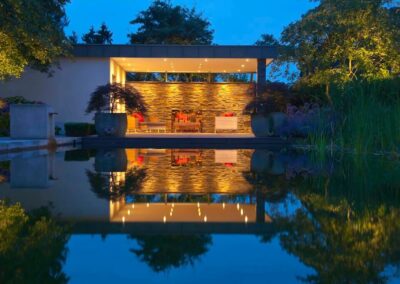 Beleuchteter Loungebereich mit Steinwand und Reflexion im Schwimmteich bei Nacht.