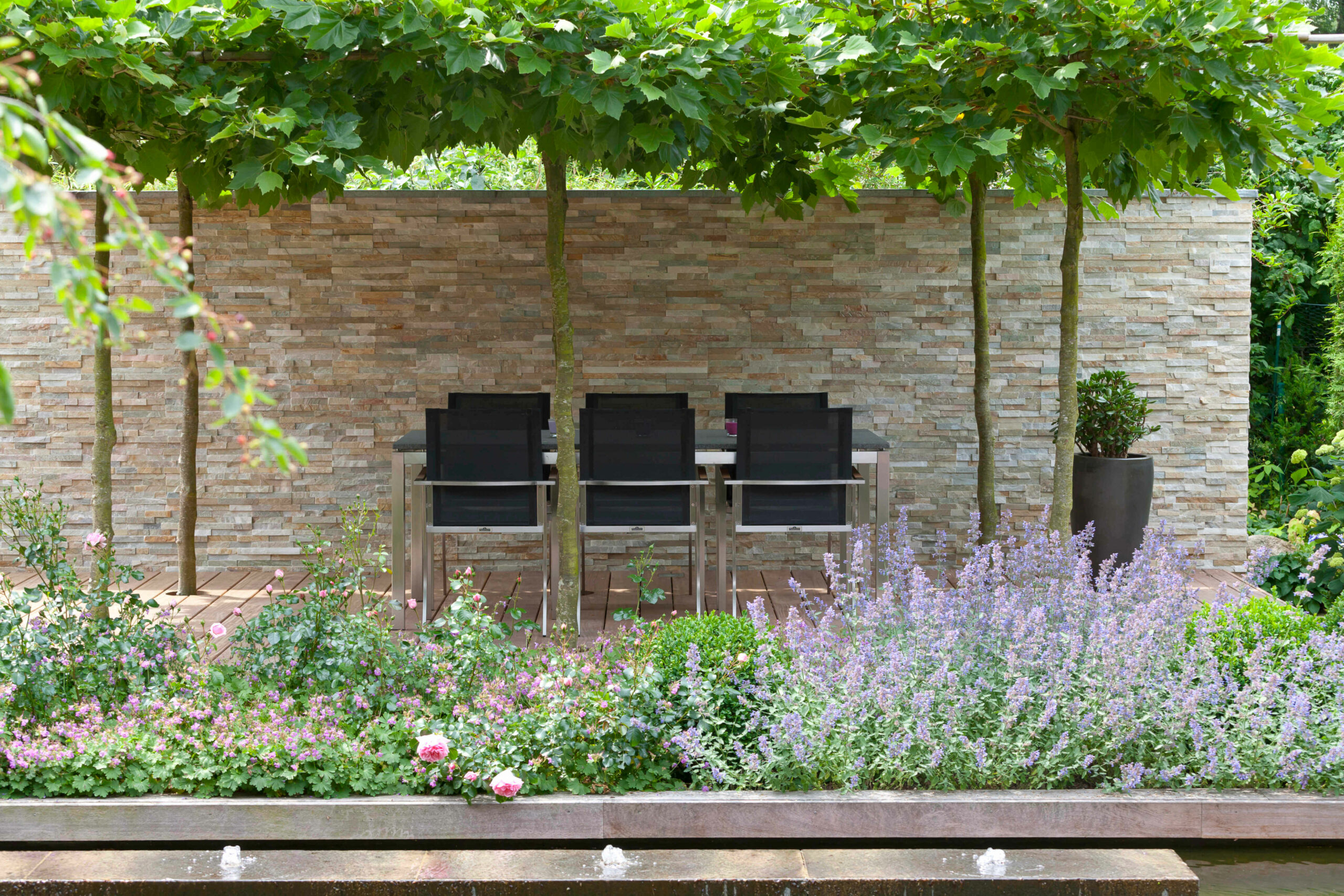 Sitzbereich im Garten mit Pflanzen und Wand Sitzbereich mit drei schwarzen Stühlen vor einer Steinwand und bunten Blumen im Garten.