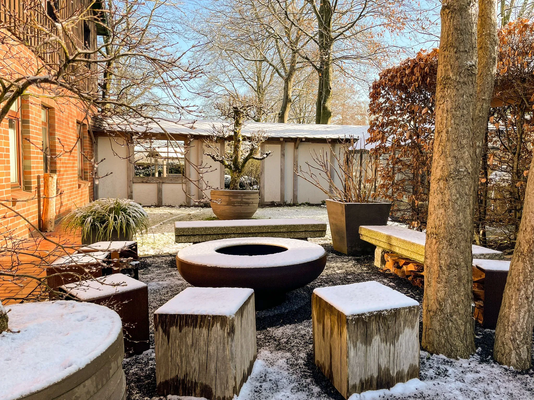 Runde Feuerstelle aus Metall in einem Garten, umgeben von Holzstühlen und schneebedeckten Pflanzen.