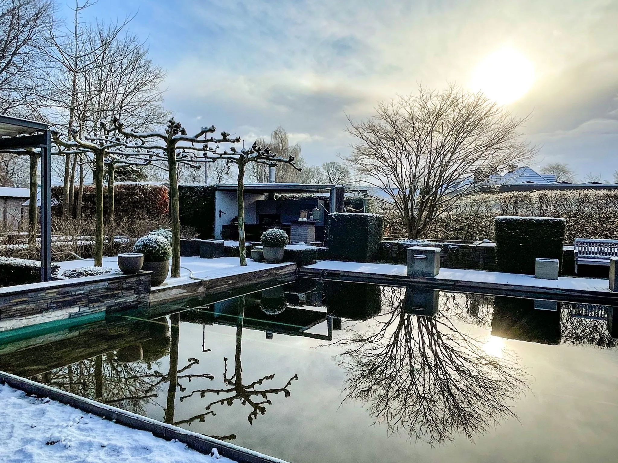 Winterlandschaft mit einem ruhigen Pool, der die Bäume und den Himmel reflektiert, umgeben von schneebedeckten Pflanzen und Strukturen.