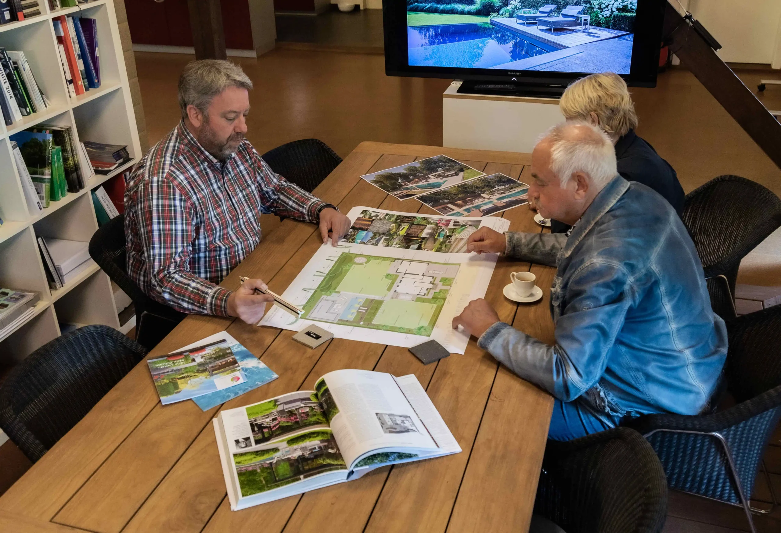 Drei Personen sitzen an einem Tisch und betrachten Pläne und Broschüren für Garten- und Landschaftsgestaltung.