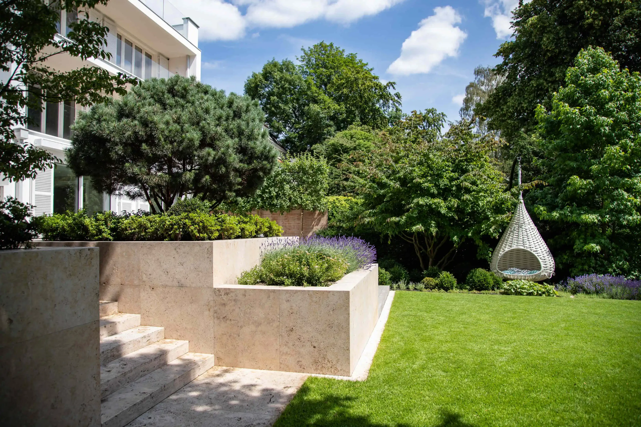 Garten mit einer modernen Steinmauer, Treppe und einer hängenden Korbschaukel, umgeben von Bäumen und Sträuchern.