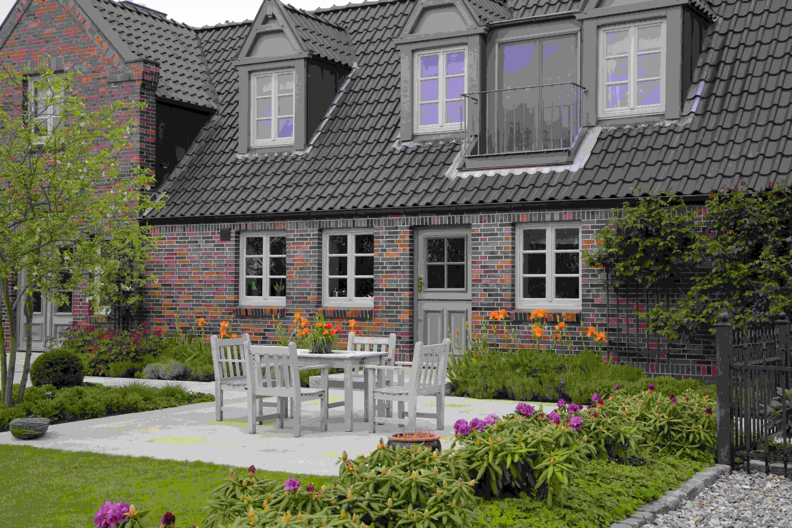 Zweistöckiges Haus mit roter Ziegelfassade und mehreren Fenstern, umgeben von einem gepflegten Garten mit Blumen und einer Terrasse mit Tisch und Stühlen.