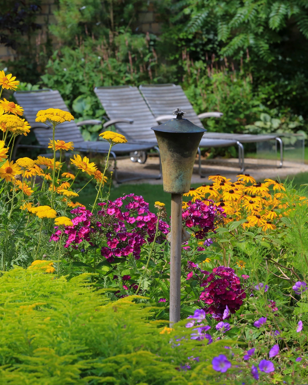 Blumenbeet mit gelben Blüten, pinken Blütenständen und violetten Blüten, zentrale Metall-Lampe, dahinter Lounge-Möbel und grüne Hecken im Hintergrund