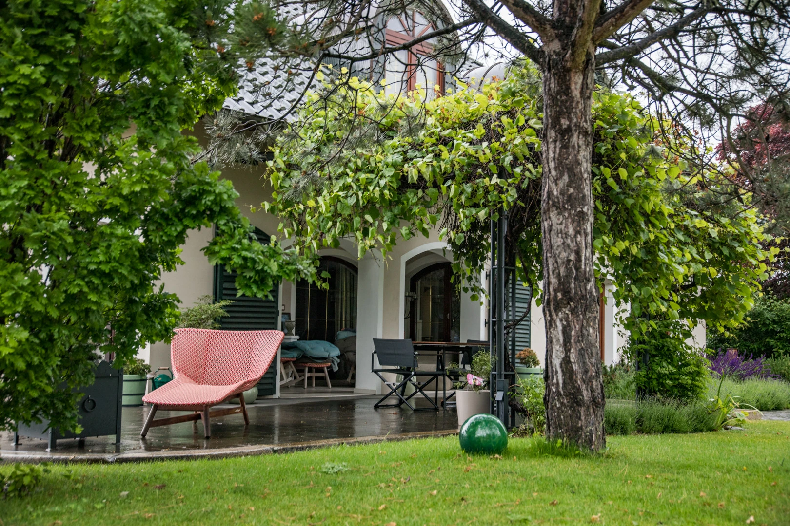Ein mit Kletterpflanzen bewachsener Pergola-Bereich liegt vor einer Villa mit Terrasse, nassem Boden und Gartenmöbeln in einem gepflegten Garten.