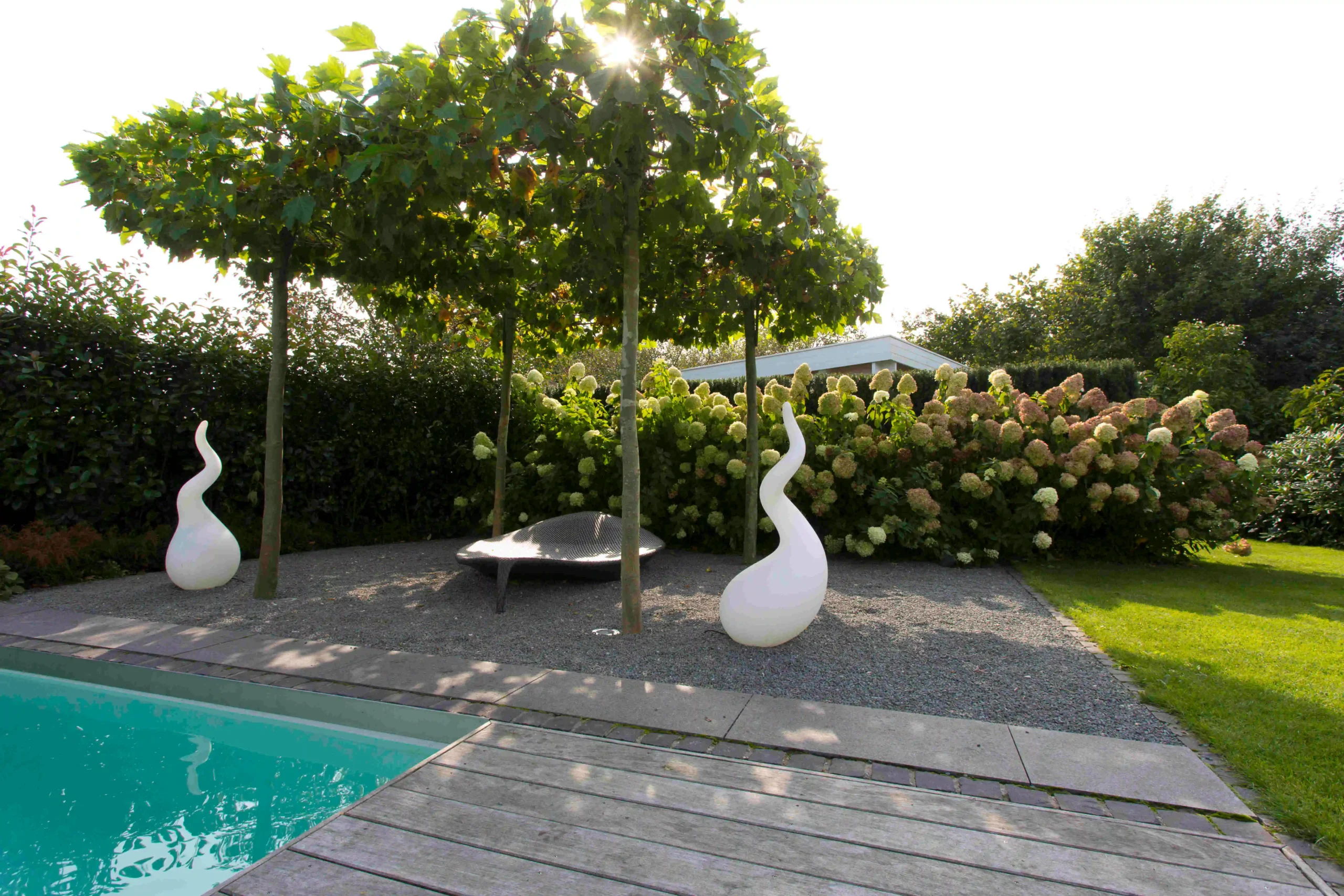 Unter einem dichten Platanendach liegen ein Naturpool, weiße Skulpturen und ein Kiesgarten vor Hortensien und Holzdeck im Sonnenlicht.