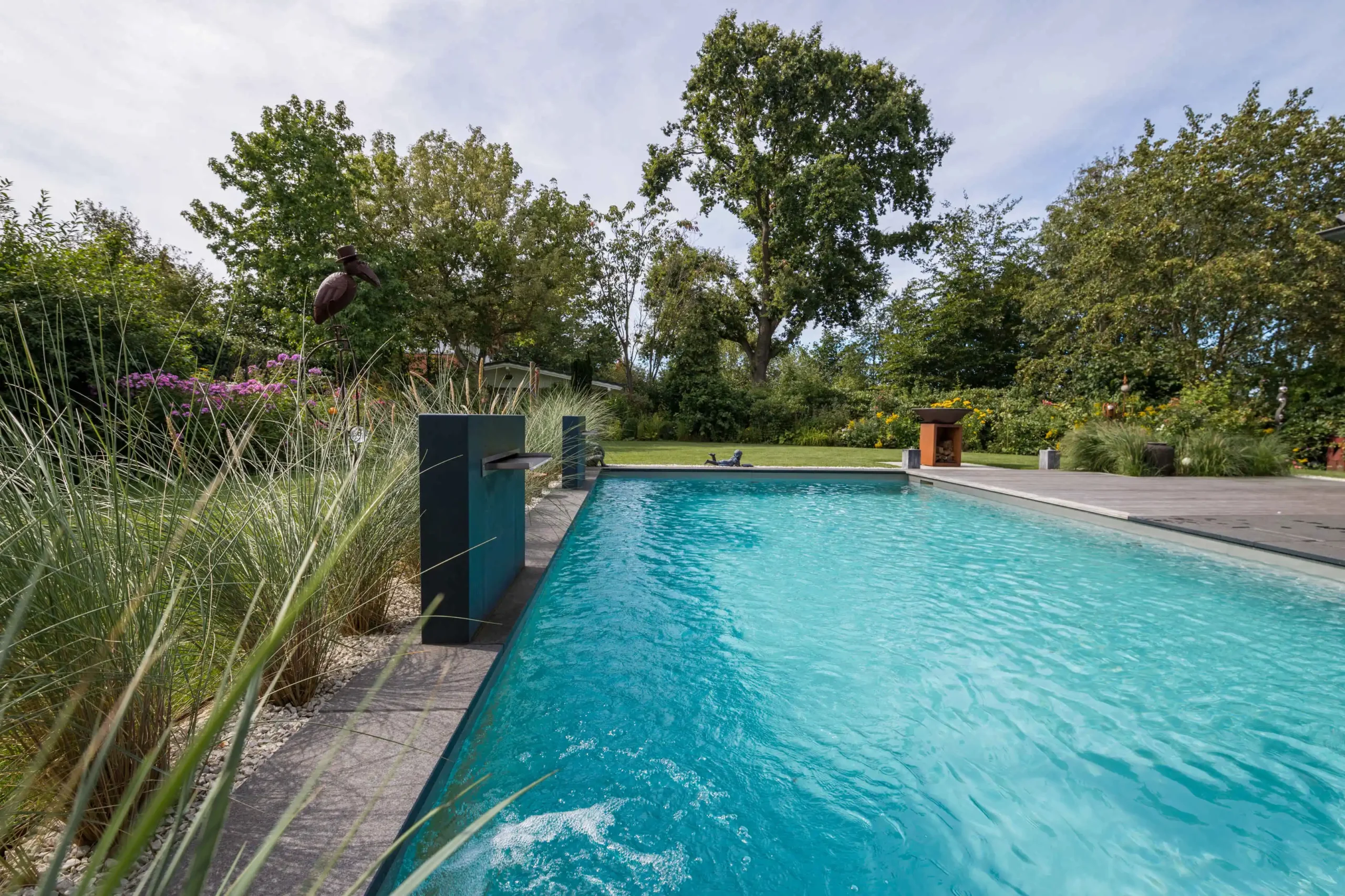 Ein türkisblauer Naturpool liegt in einem üppig bepflanzten Garten mit Holzdeck und hohen Bäumen.