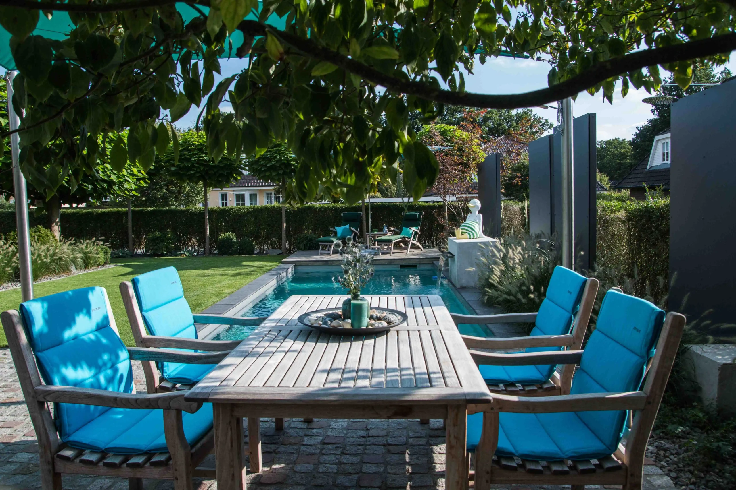Ein rechteckiger Naturpool liegt in einem gepflegten Garten mit Liegen, Holzdeck und türkisfarbenen Polstern.
