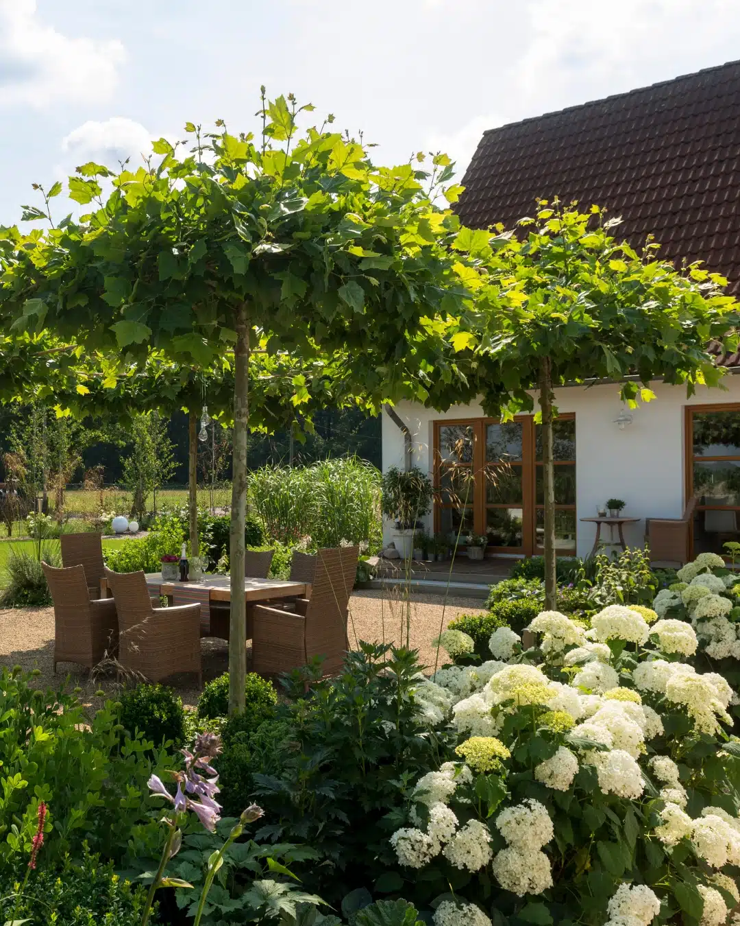 Sonniger Sitzbereich mit Holztisch und Korbstühlen unter begrünten Bäumen, umgeben von üppigen Beeten mit weißen Blüten.