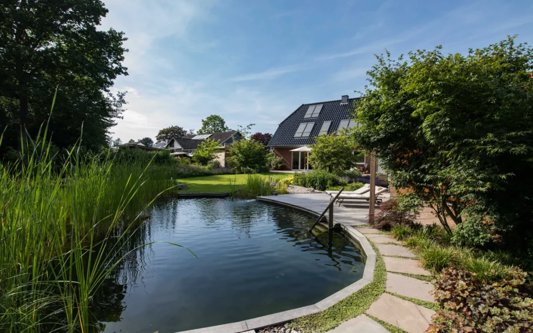 Ein naturnaher Schwimmteich liegt im Garten eines Hauses mit Holzdeck, Liegen und dichter Bepflanzung.