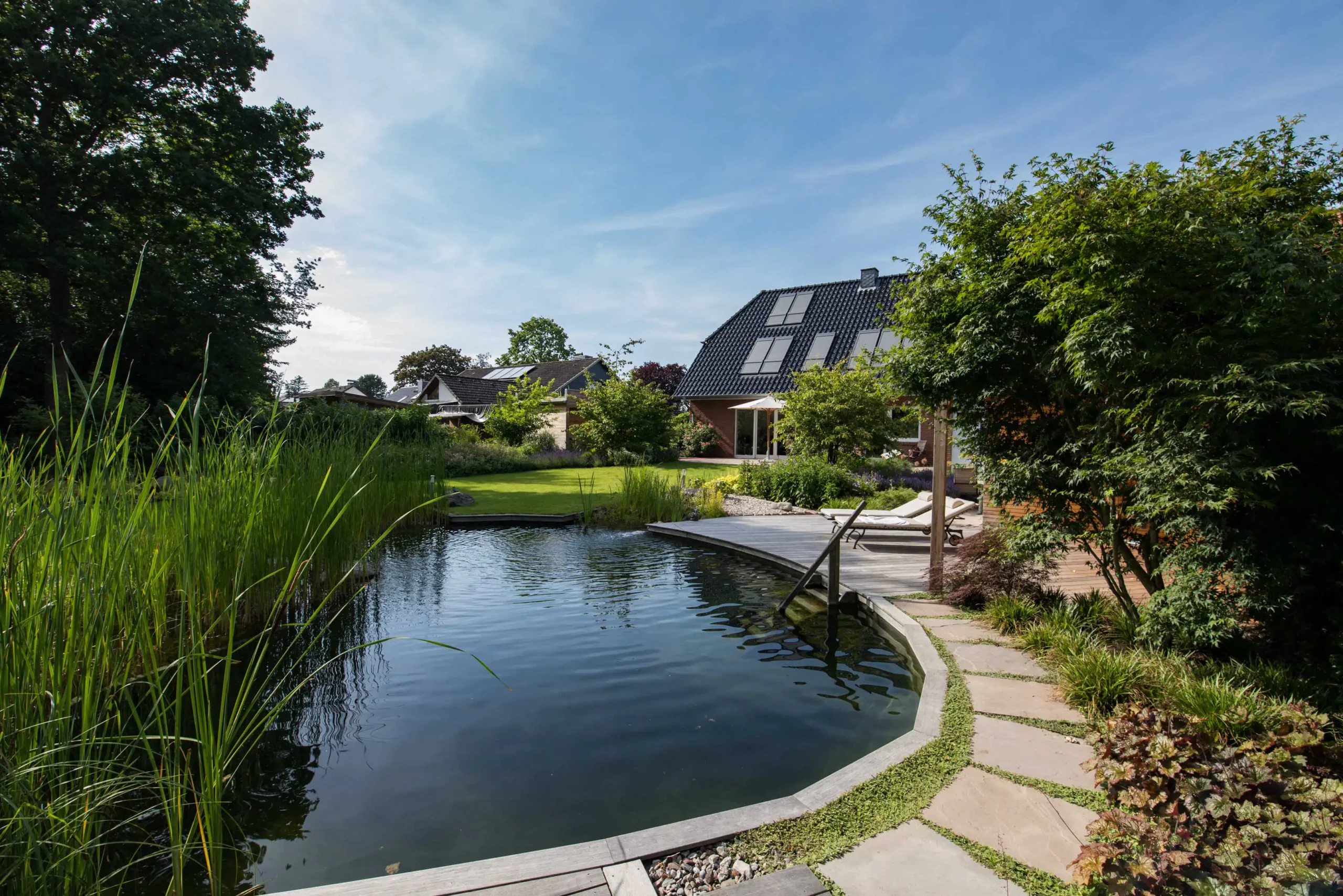 Ein naturnaher Schwimmteich liegt im Garten eines Hauses mit Holzdeck, Liegen und dichter Bepflanzung.