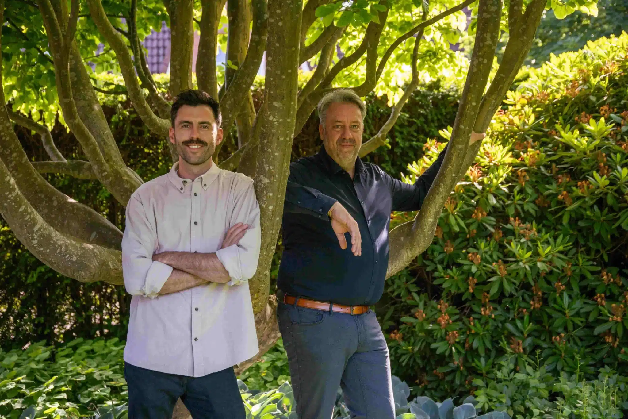 Im grünen Garten stehen Torben Stuewe und Christian Bahl entspannt unter einem ausladenden Baum mit dichtem Blattwerk.