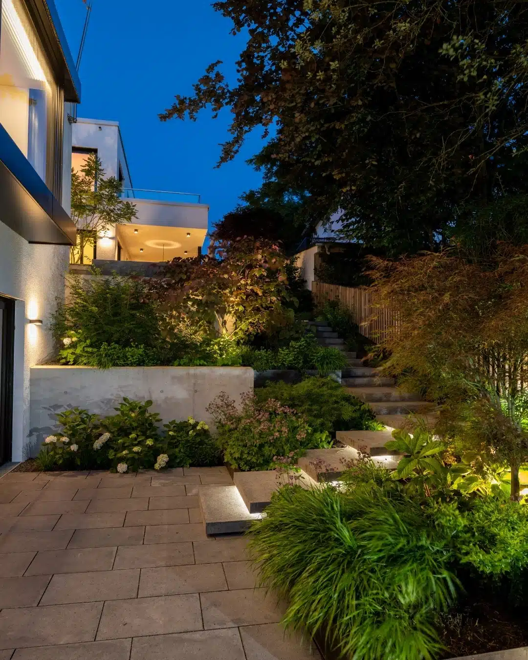 Beleuchteter Garten mit bepflanzten Terrassen, Treppenstufen und modernen Wohnhäusern bei einbrechender Dämmerung