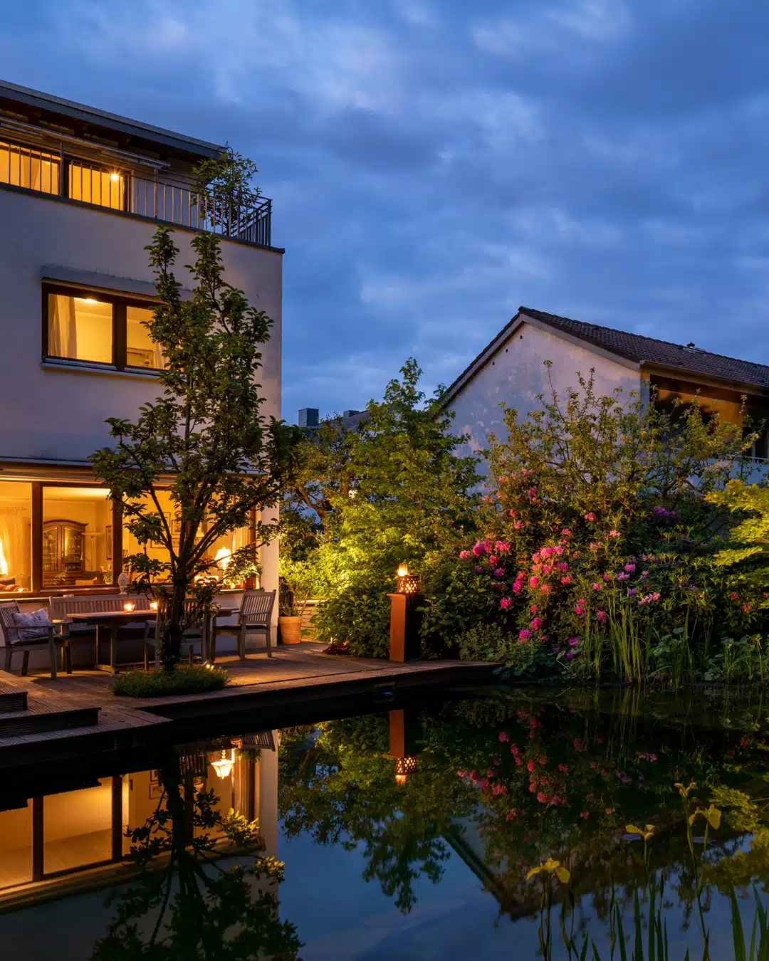 Beleuchteter Garten mit blühenden Sträuchern, Sitzplatz und Teich neben Wohnhäusern in der Abenddämmerung