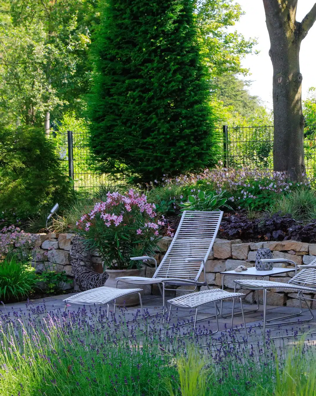 Gepflegter Gartenbereich mit Liegestühlen, blühenden Stauden, Lavendel und einer Natursteinmauer unter altem Baumbestand.