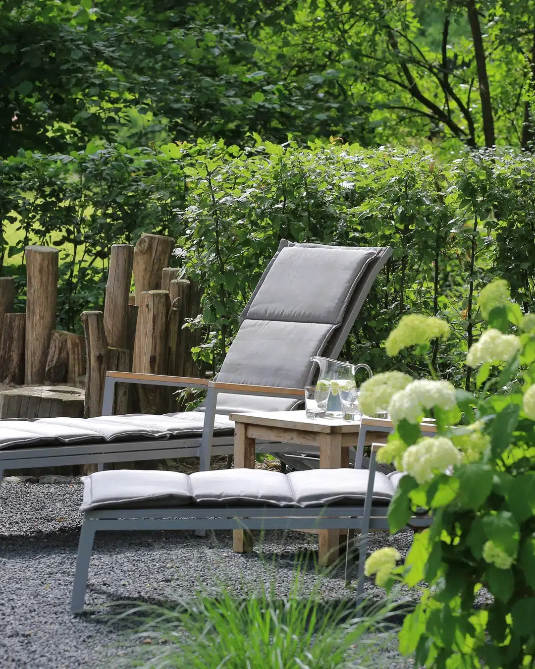 Graue Liegestühle und ein Beistelltisch mit Gläsern stehen in einem grünen Garten neben einer blühenden Hortensie.