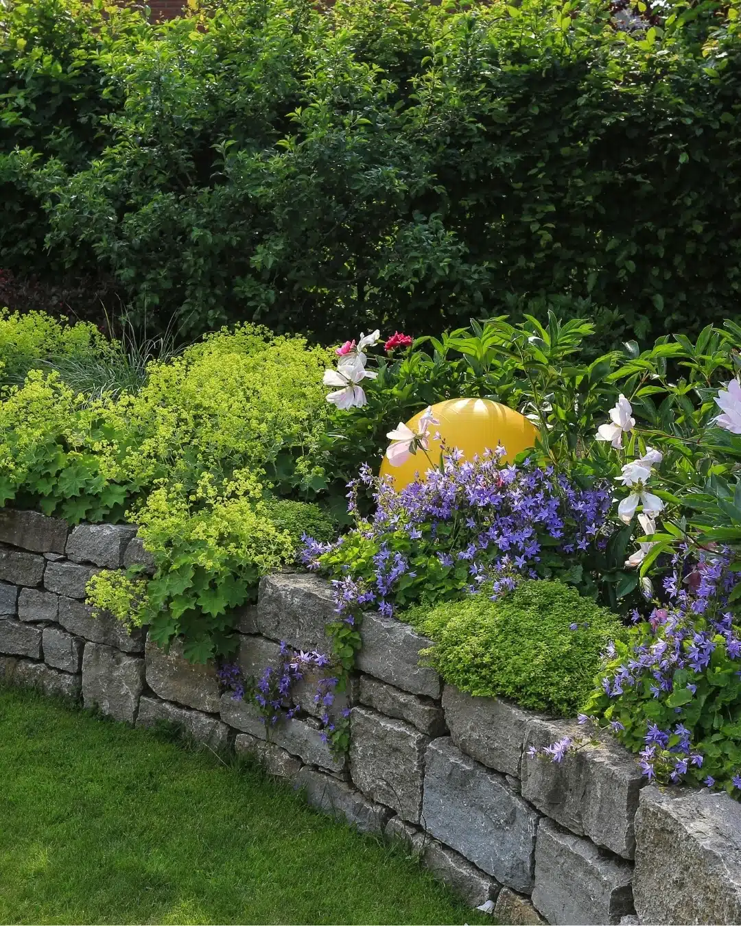 Bepflanzte Natursteinmauer mit üppigen Stauden, violetten Blüten, grünem Laub und einer gelben Gartenlampe