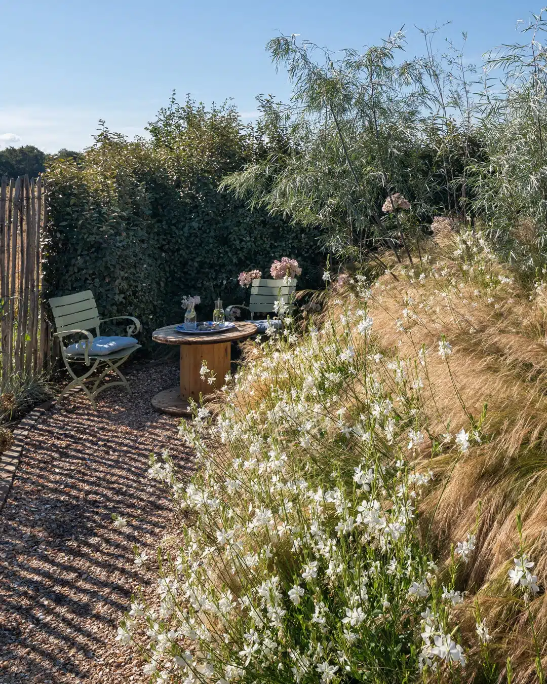 In einem üppig bepflanzten Garten laden zwei Stühle und ein gedeckter runder Tisch zwischen Stauden mit weißen Blüten und Gräsern zum Verweilen ein.