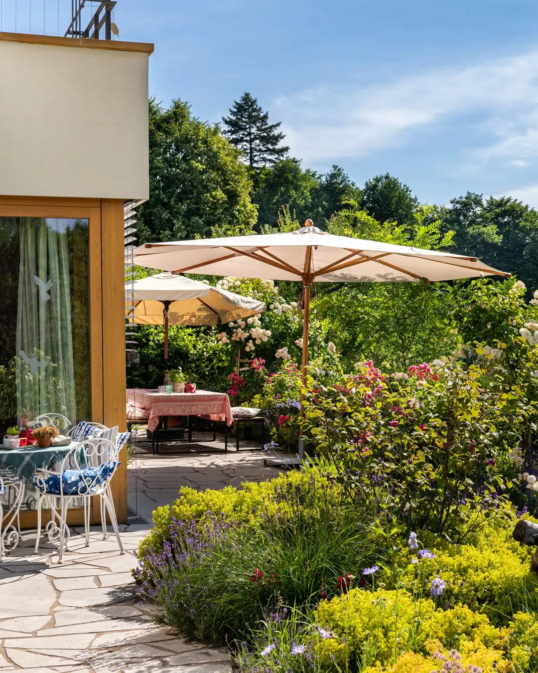 Gepflegte Gartenterrasse mit großen Sonnenschirmen, Sitzplätzen, blühenden Rosen und üppiger Bepflanzung im Grünen.